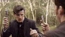A young man in a suit, holding a sonic screwdriver, looks intently at another person who is mirroring him in a lush green forest setting.