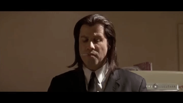 A man in a black suit with long hair, wearing a white shirt and tie, stands with a cigarette in his mouth and eyes closed, set against an indoor background.